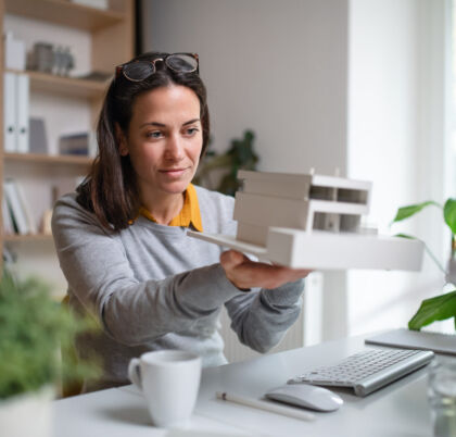 Architect With Model Of A House Sitting At The Desk Indoors In Office.