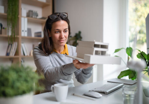 Architect With Model Of A House Sitting At The Desk Indoors In Office.